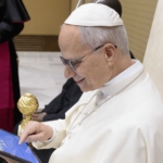 Papa Leão sentado, sorrindo enquanto utiliza um tablet durante um encontro no Vaticano