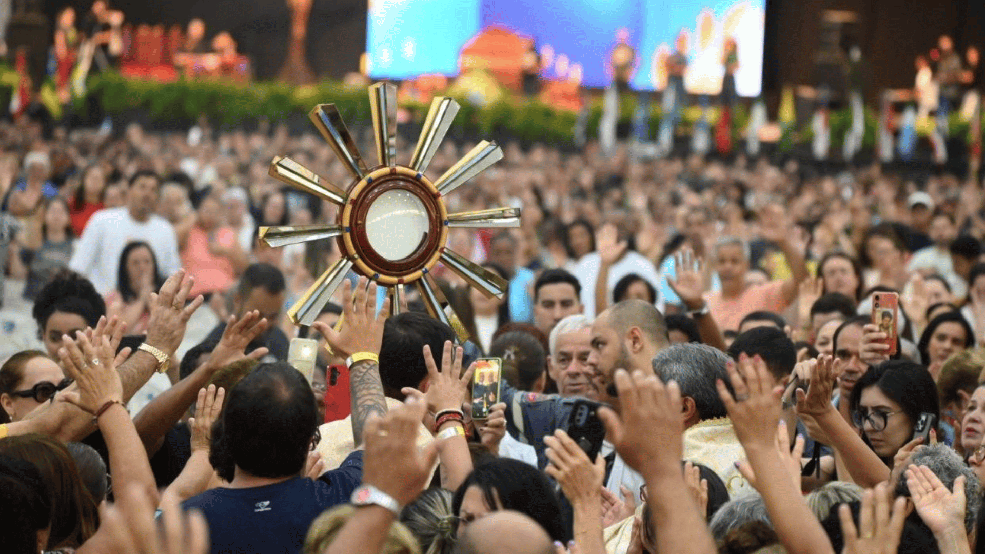 A imagem ilustra um ostensório dourado com a hóstia consagrada sendo conduzido por um sacerdote em meio a alguns peregrinos que estão com as mãos estendidas para o alto.