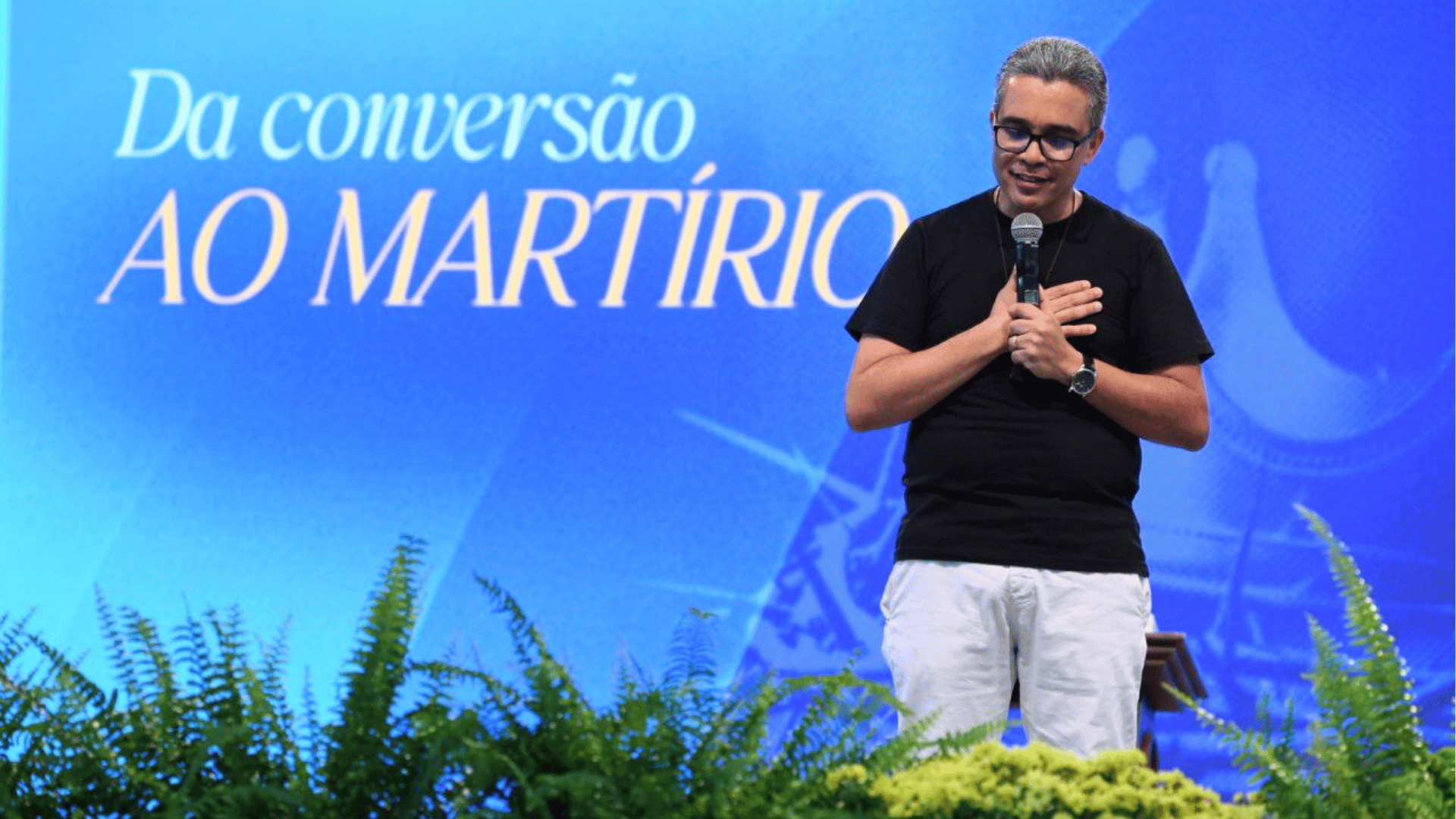 Alexandre Oliveira pregou sobre o tema da conversão Alexandre Oliveira aparece em pé, com camiseta preta, calça branca, segura um microfone e coloca as mãos sobre o peito. Um telão atrás dele exibe o escrito "Da conversão ao martírio", tema de sua pregação no Hosana Brasil 2025.