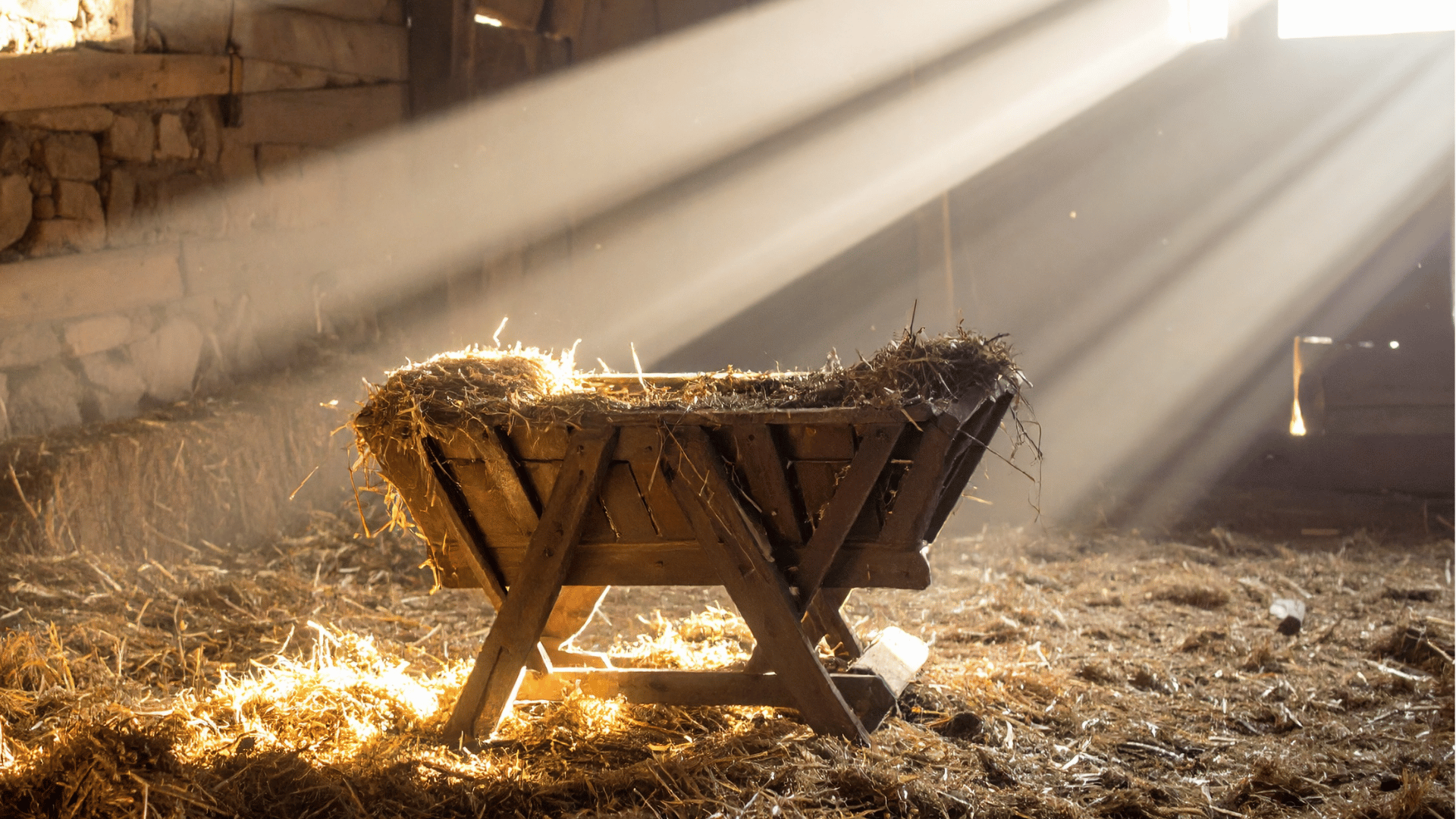 Uma manjedoura aparece em um chão coberto por palha, sendo iluminada por raios de luz vindos de uma janela