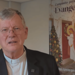 o cardeal jaime spengler aparece com óculos, camisa branca e terno preto. Ao fundo, o cartaz da campanha para a evangelização 2025