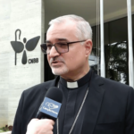 Dom João Justino fala ao microfone do Jornalismo Canção Nova. Ao seu fundo, aparece o prédio que é a sede da CNBB em Brasília