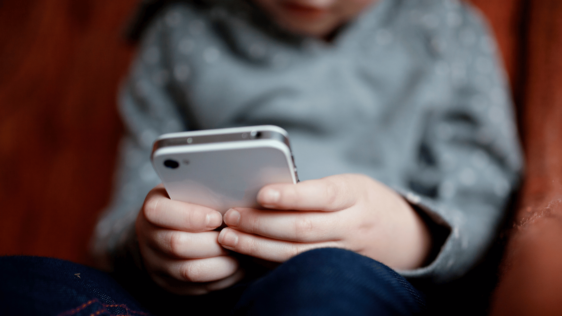 A imagem ilustra uma criança com uma camisa de manga longa cinza com um celular branco nas mãos.