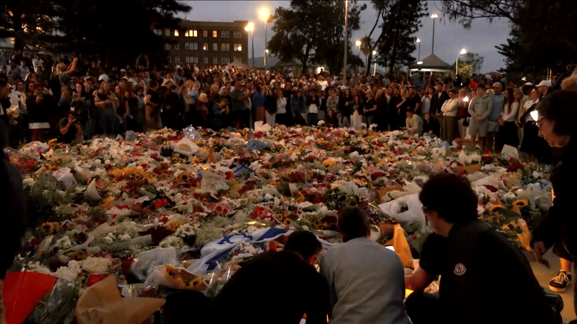 Homenagem a vítimas do atentado terrorista em Sidney A imagem ilustra uma multidão ao redor de uma área tomada por flores, velas e outros objetos em homenagem às vítimas de um ataque terrorista que aconteceu no local.