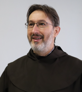 A imagem ilustra um religioso carmelita, usando hábito religioso. É um homem branco, de barba grisalha e cabelo castanho liso.