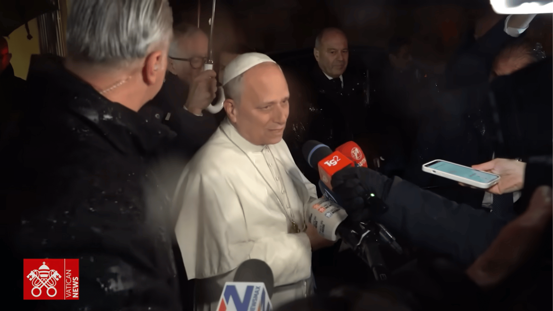 O Papa Leão XIV aparece com suas habituais vestes brancas rodeado por jornalistas com microfones e câmeras