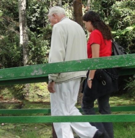 Uma mulher - secretária do padre Jonas Abib - de camiseta vermelha e calça escura, segurando uma bolsa, caminha ao lado do padre Jonas Abib, que usa calça branca e um casaco bege