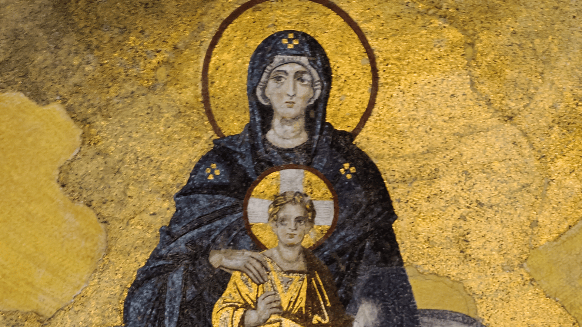 A imagem ilustra um mosaico da Virgem Maria, representada como uma mulher de pele branca e vestes azul escuro, e o Menino Jesus, representado como uma criança de pele branca e vestes douradas. A mão direita da Virgem Maria encontra-se sobre o ombro direito do Menino Jesus.