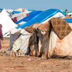Mulher vestindo roupas tradicionais caminha entre barracas improvisadas de um campo de refugiados, com lonas azuis e brancas, chão árido e objetos domésticos visíveis ao redor.
