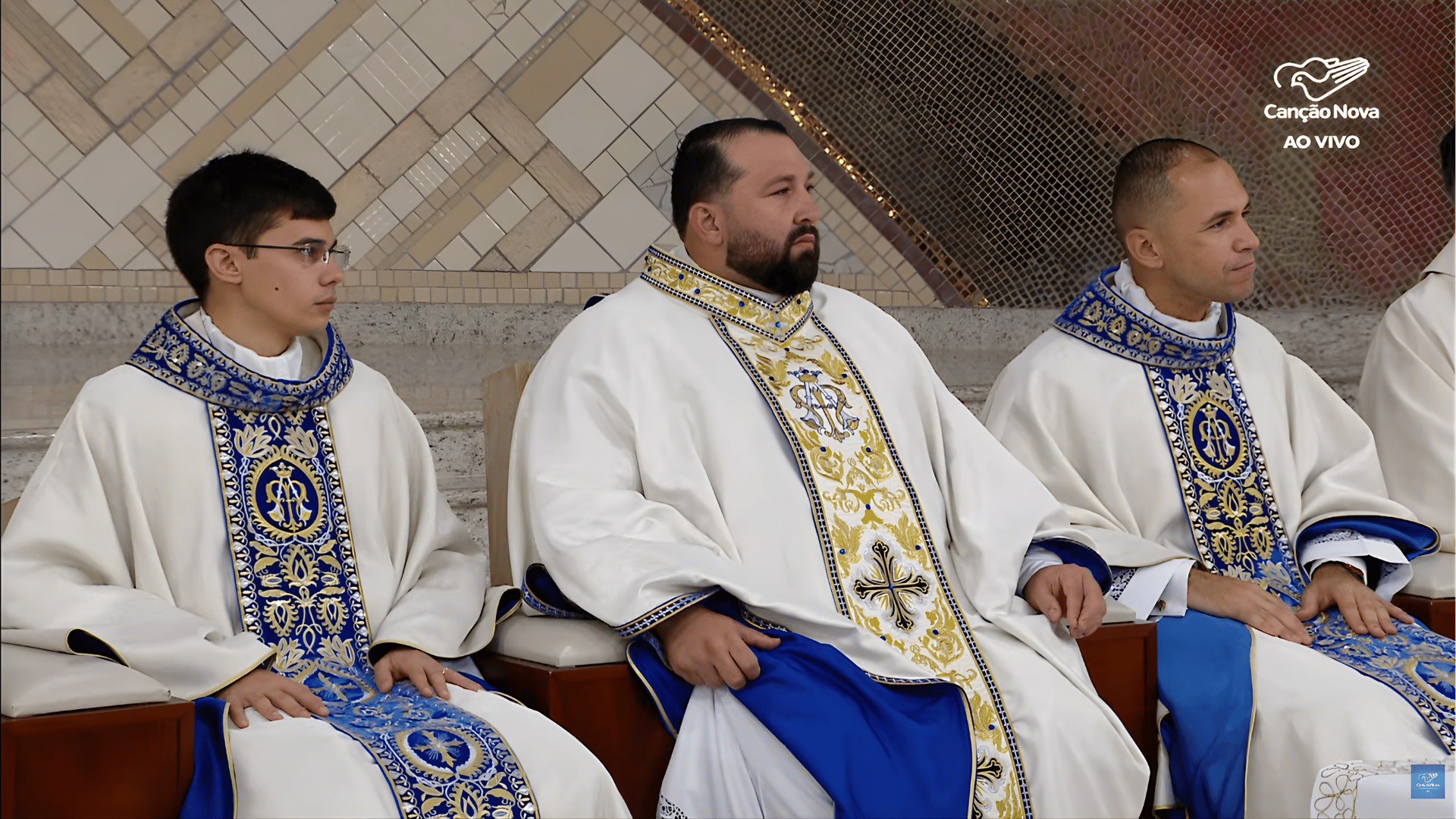 Sacerdotes sentados no presbitério durante a celebração, com paramentos litúrgicos em azul e dourado