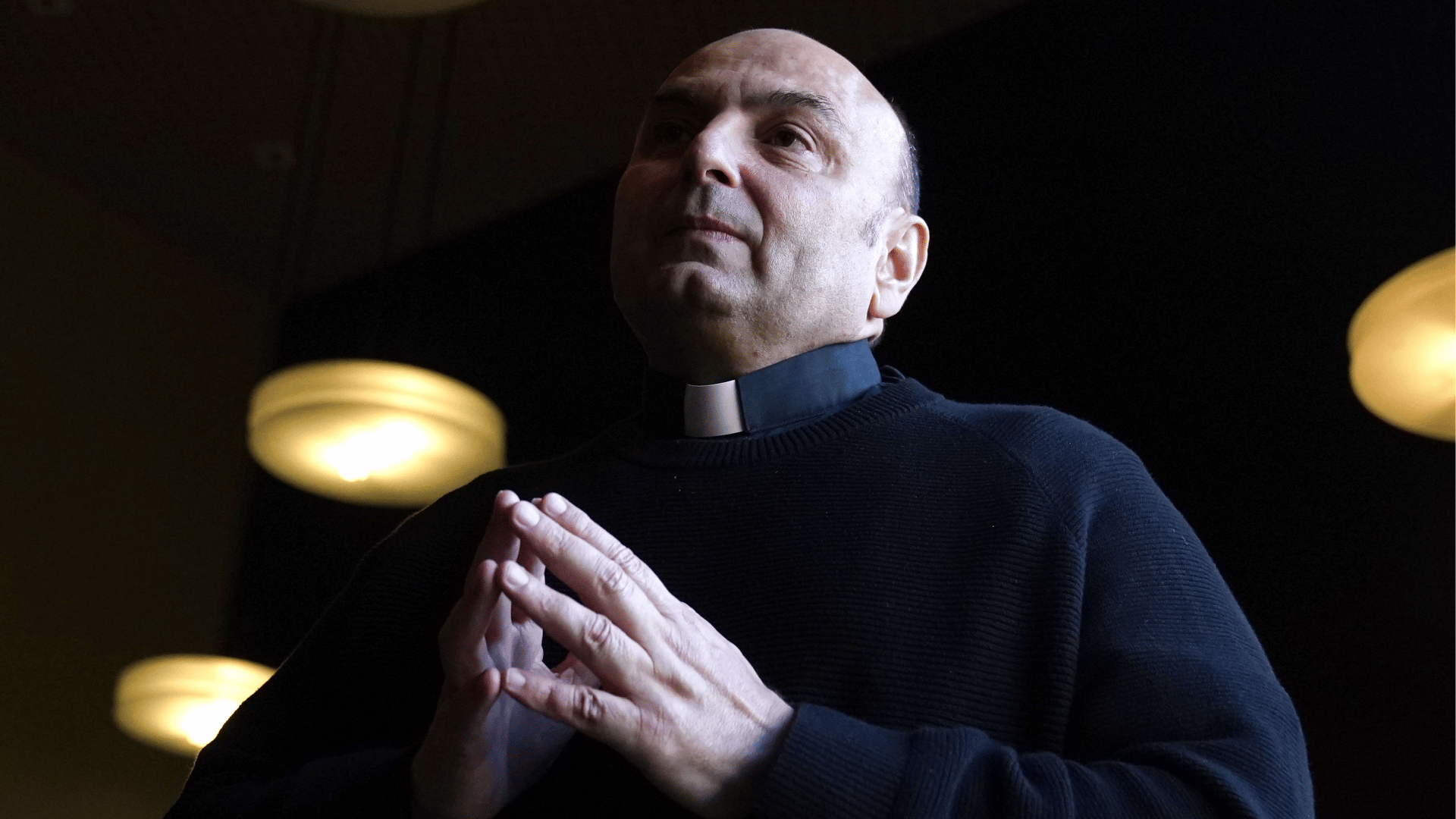 Sacerdote com as mãos postas em atitude de oração, iluminado por luz suave no interior de uma igreja