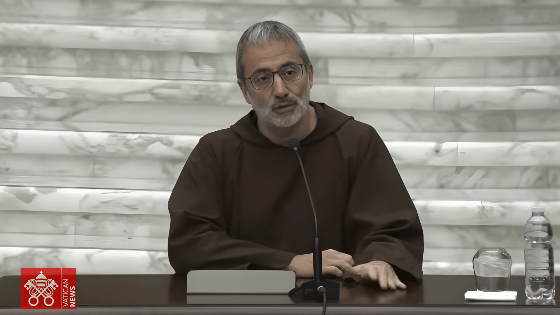 O padre Roberto Pasolini aparece sentado à frente de uma mesa falando ao microfone