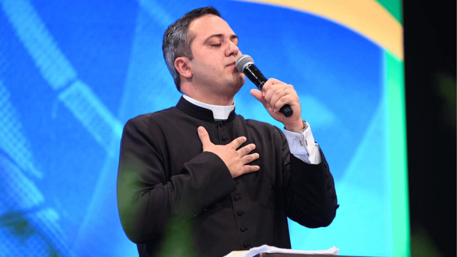 Um homem aparece em pé, com olhos fechados, uma das mãos no peito e a outra segurando um microfone. O homem é o padre Sydnei Dias, que está de batina preta.