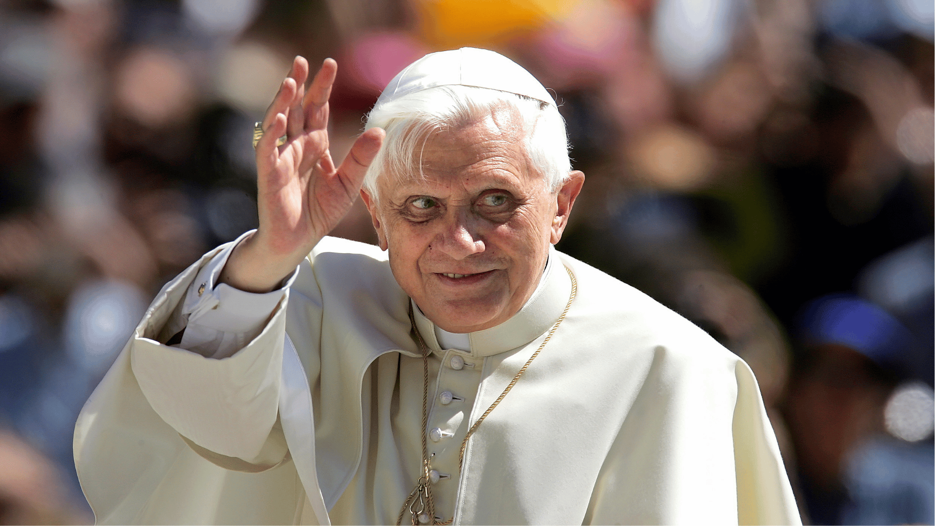 Papa Bento XVI acena aos fiéis durante celebração ao ar livre, vestindo batina branca e solidéu, diante de uma multidão.
