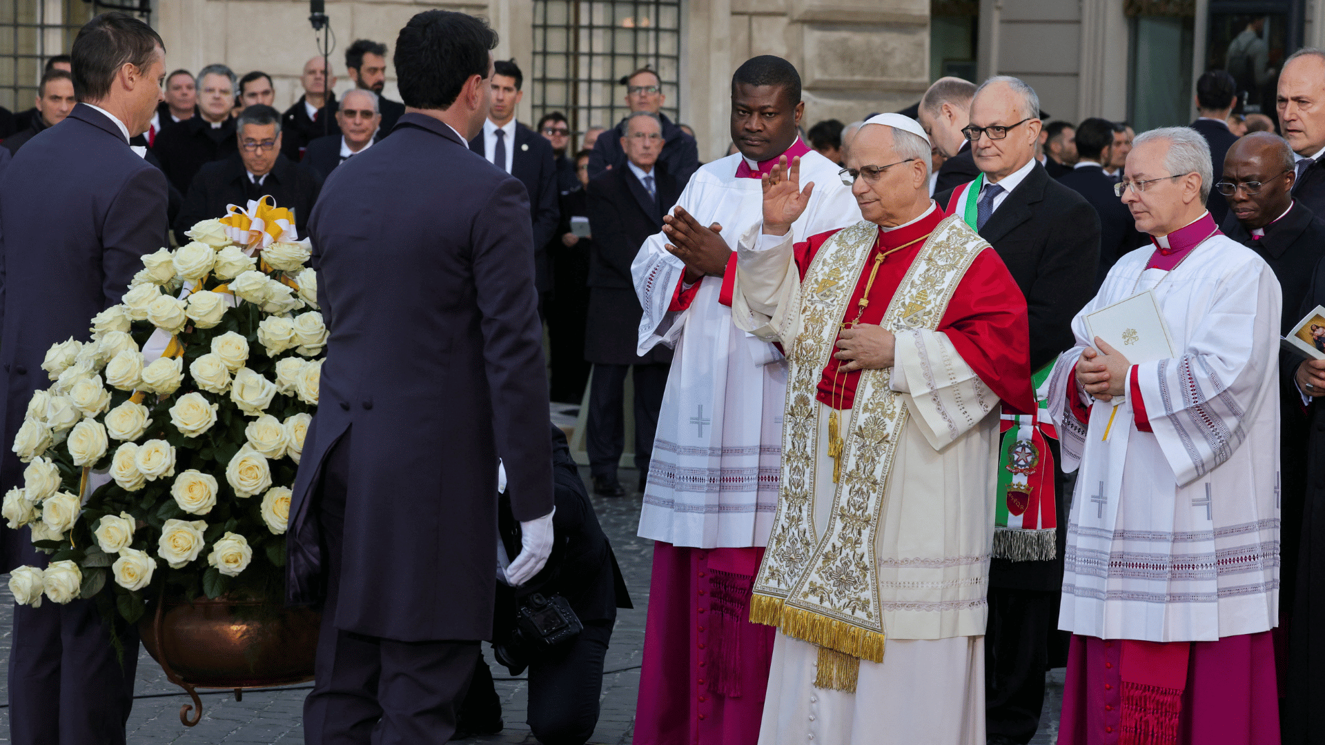 A imagem ilustra o Papa Leão XIV usando uma estola e com a mão erguida enquanto abençoa uma coroa de flores que foi ofertada a Nossa Senhora. O Pontífice encontra-se cercado por outros religiosos e pelo prefeito de Roma.