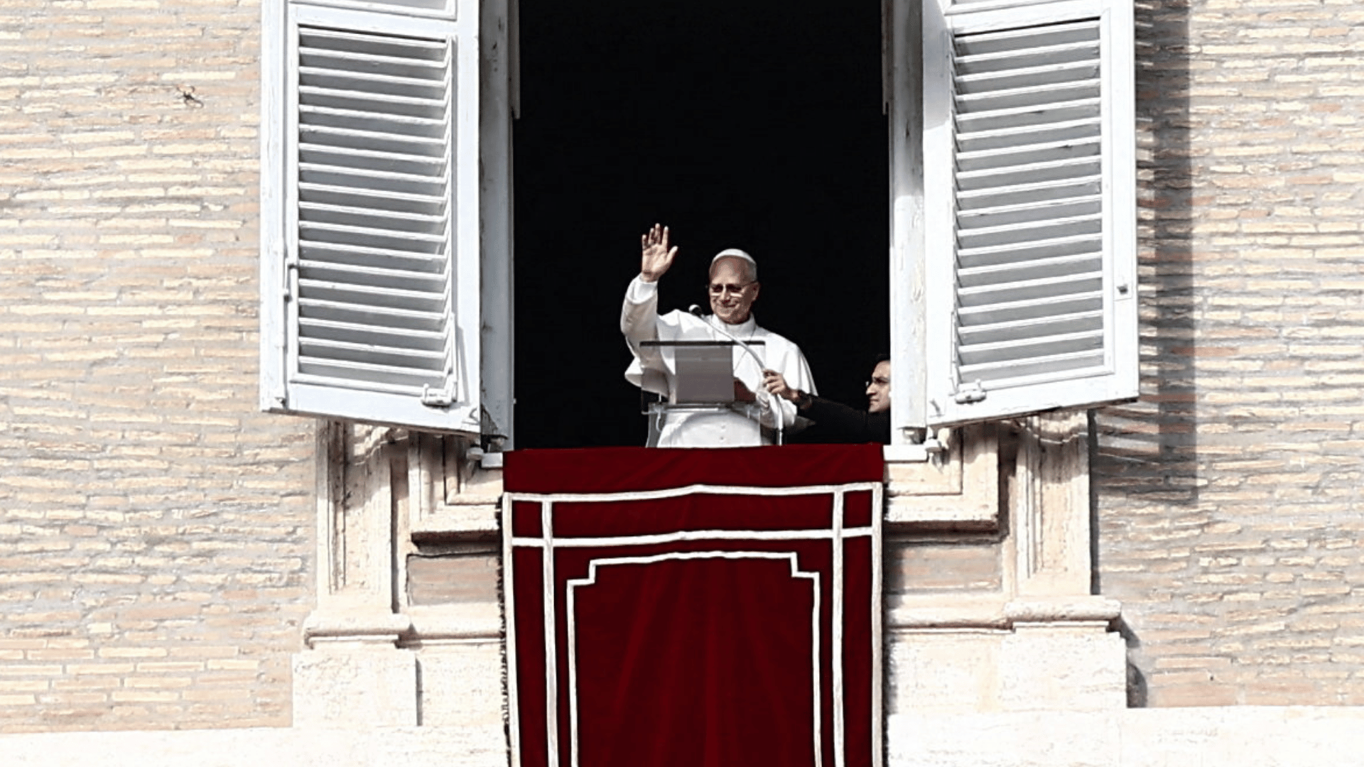 A imagem ilustra o Papa Leão XIV em suas tradicionais vestes pontifícias brancas, coma mão direita erguida em gesto de aceno para fiéis. Ele encontra-se diante do parapeito de uma das janelas do Palácio Apostólico, no Vaticano.