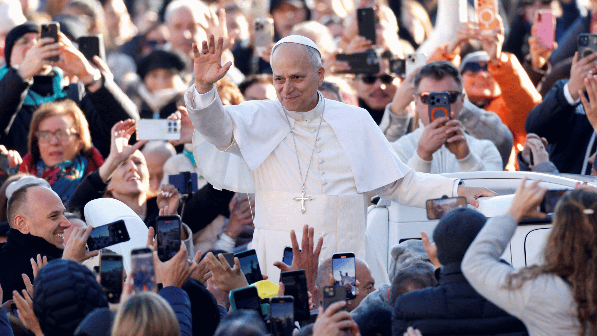 Papa Leão XIV acena para fiéis durante "giro do papamóvel" A imagem ilustra o Papa Leão XIV em pé no papamóvel, passando pelos fiéis e acenando para eles. As pessoas ao redor do Pontífice seguram celulares para filmá-lo e acenam de volta.