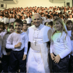 A imagem mostra o Papa Leão XIV com suas habituais vestes brancas abraçando duas crianças. Várias outras crianças estão atrás deles.