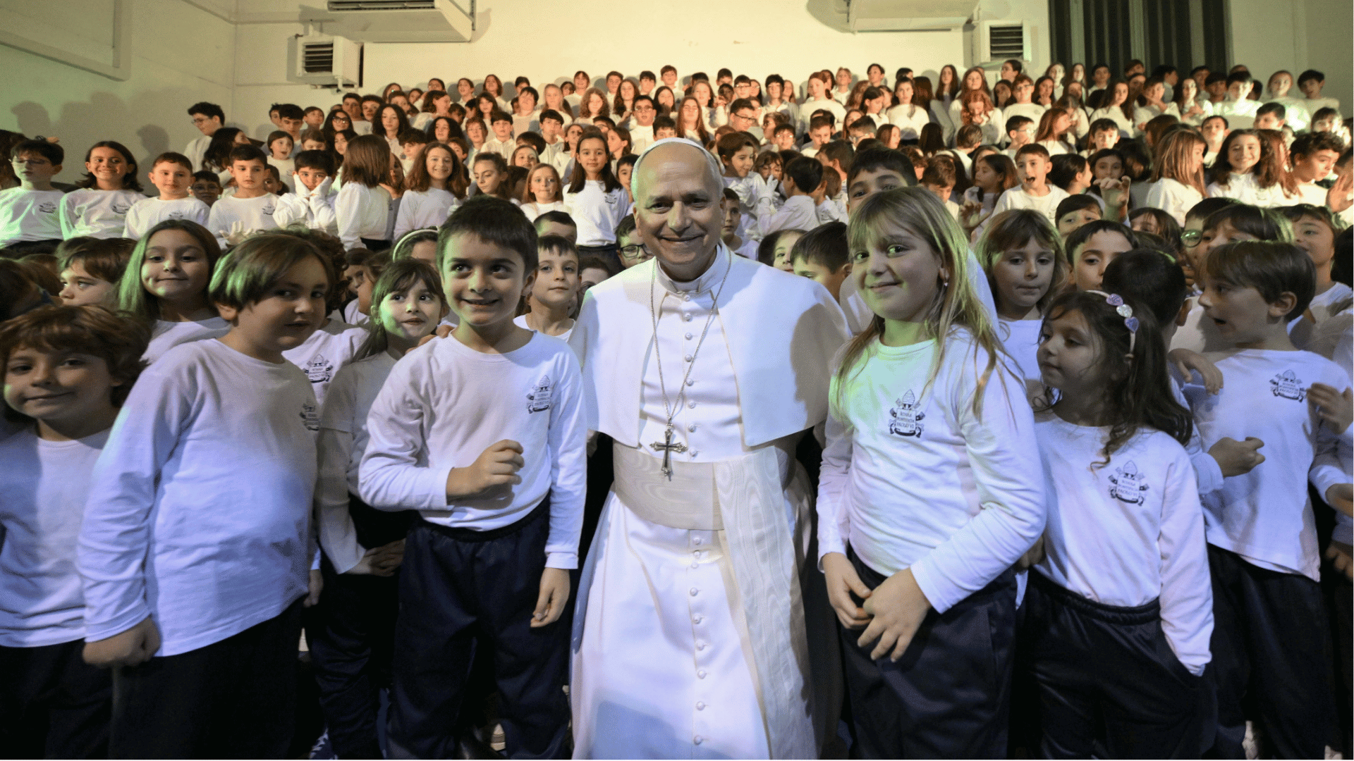 A imagem mostra o Papa Leão XIV com suas habituais vestes brancas abraçando duas crianças. Várias outras crianças estão atrás deles.