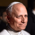 Papa sorrindo durante uma aparição pública noturna, com pessoas ao fundo