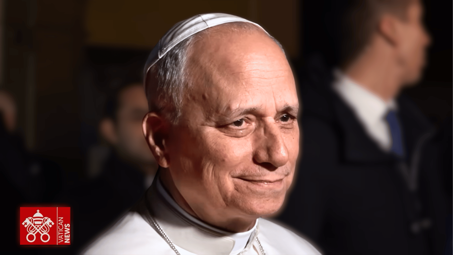 Papa sorrindo durante uma aparição pública noturna, com pessoas ao fundo