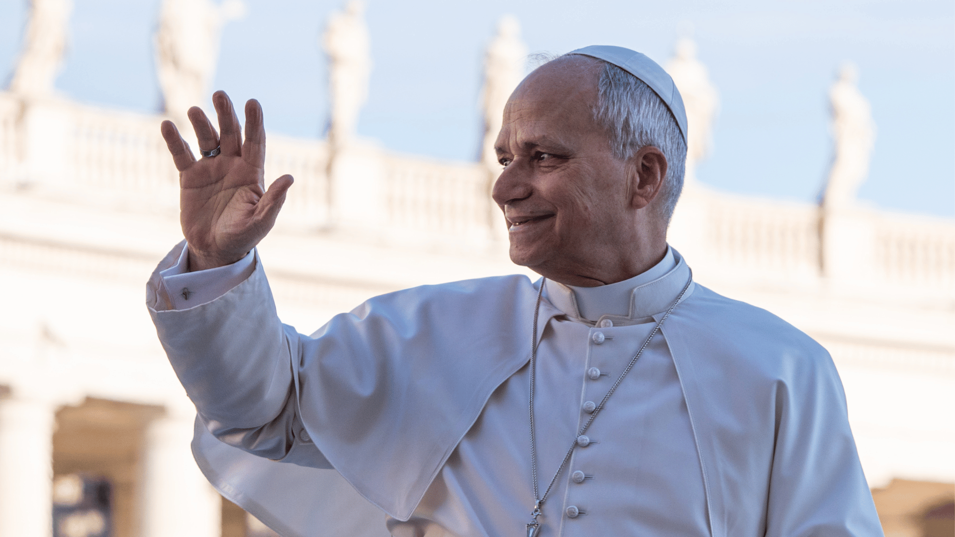 A imagem ilustra o Papa Leão XIV com suas habituais vestes pontifícias. Ele está de lado, acenando com a mão direita e com um sorriso no rosto.
