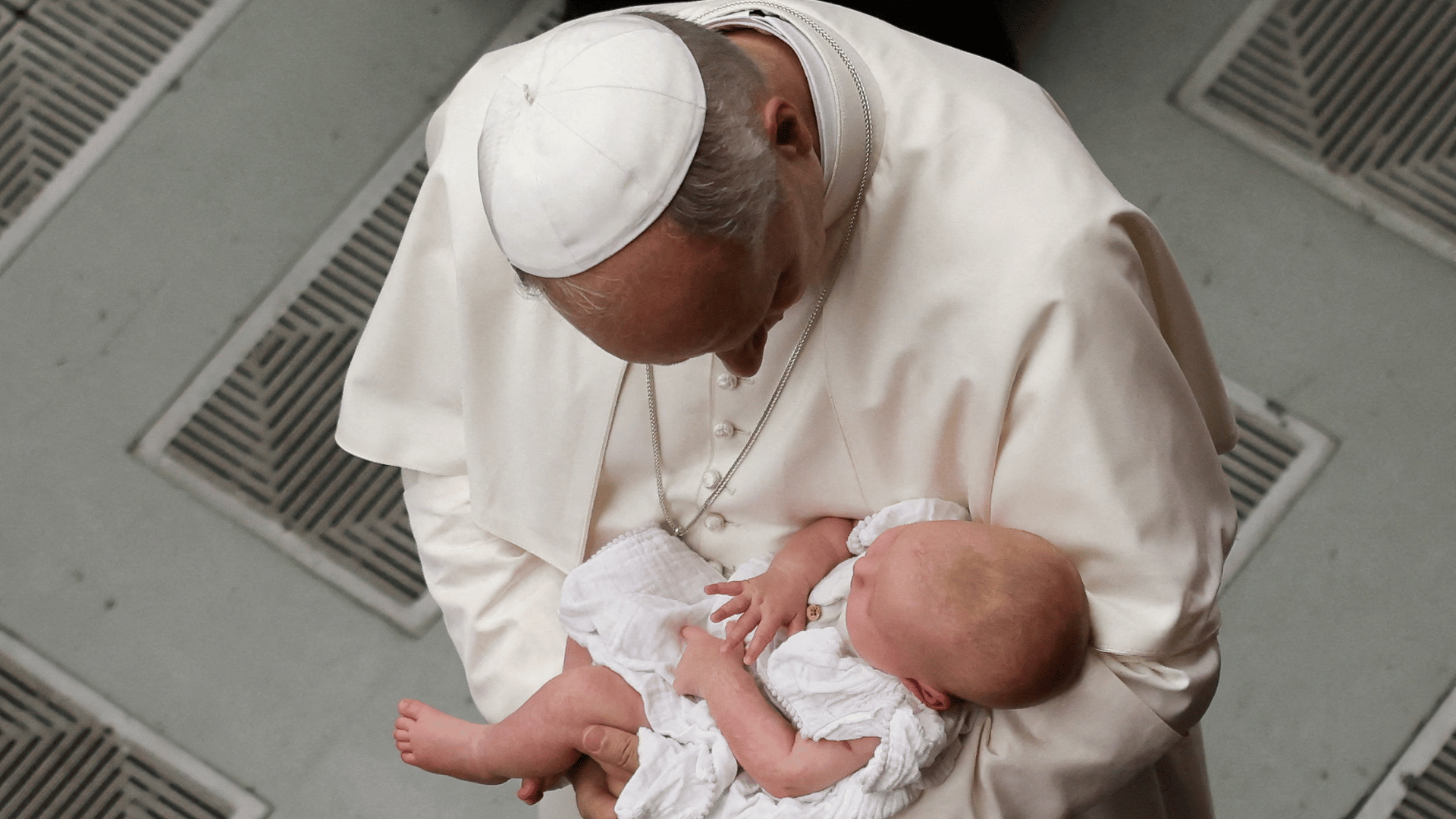A imagem ilustra o Papa Leão XIV visto de cima, com suas habituais vestes pontifícias e um bebê com vestes brancas em seu colo.