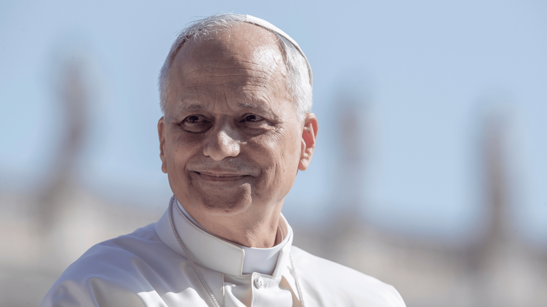 A imagem ilustra o Papa Leão XIV com suas habituais vestes pontifícias e um leve sorriso, com o olhar voltado para o lado.