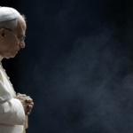 A imagem ilustra o Papa Leão XIV com suas habituais vestes pontifícias, de perfil, com as mãos unidas sobre o peito e uma expressão compenetrada.