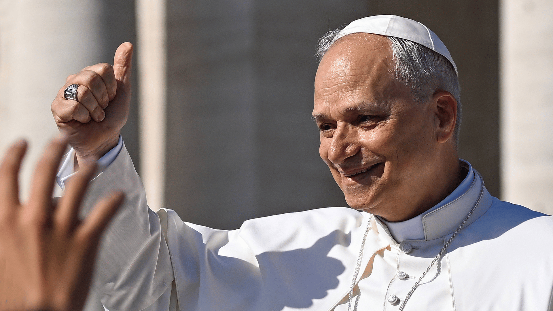 A imagem ilustra o Papa Leão XIV com suas habituais vestes pontifícias, um sorriso e a mão direita erguida com um gesto positivo.