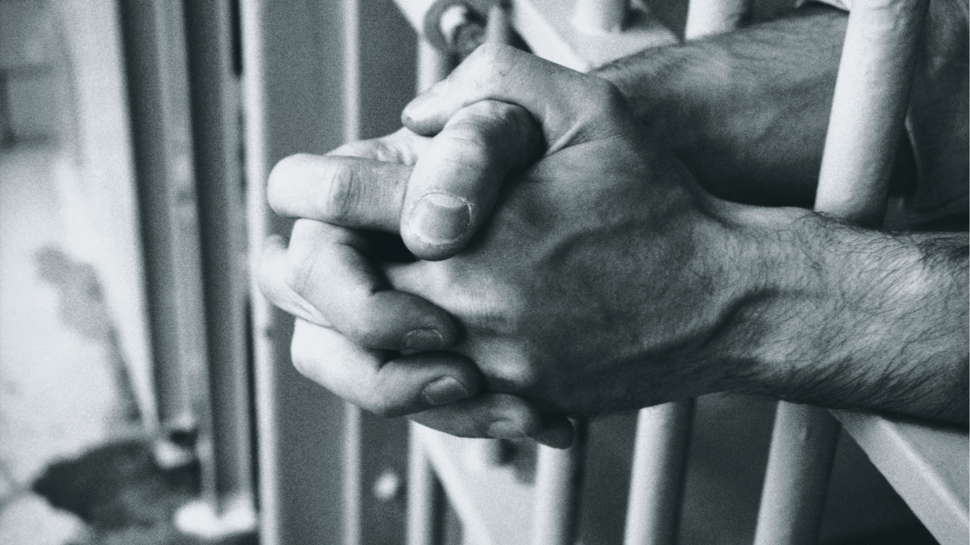 A imagem mostra as mãos de um homem unidas e apoiadas sobre as grades de uma cela