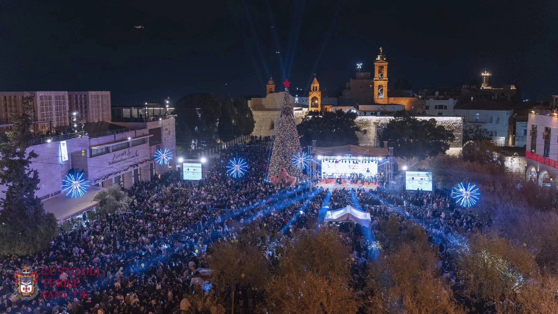A imagem mostra a Praça da Natividade, em Belém, na Terra Santa. Milhares de pessoas estão reunidas ao redor de um palco e de uma grande árvore de Natal, ambos iluminaods. 