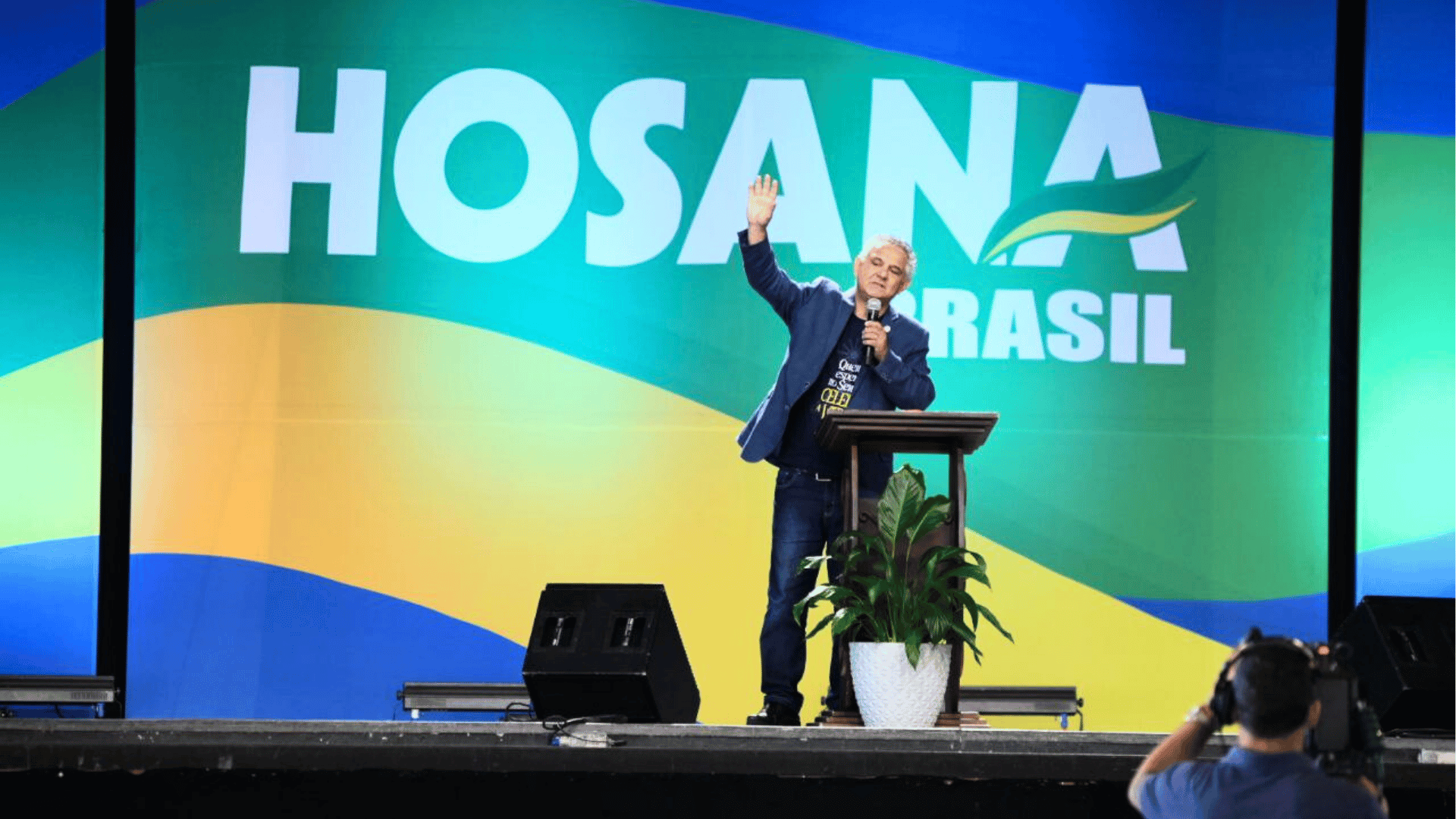 O diácono Nelsinho Corrêa durante pregação A imagem mostra um homem - o diácono Nelsinho Corrêa - em pé, segurando um microfone em uma mão, a outra está erguida para o alto. Ao fundo do palco, um telão com o escrito "Hosana Brasil"