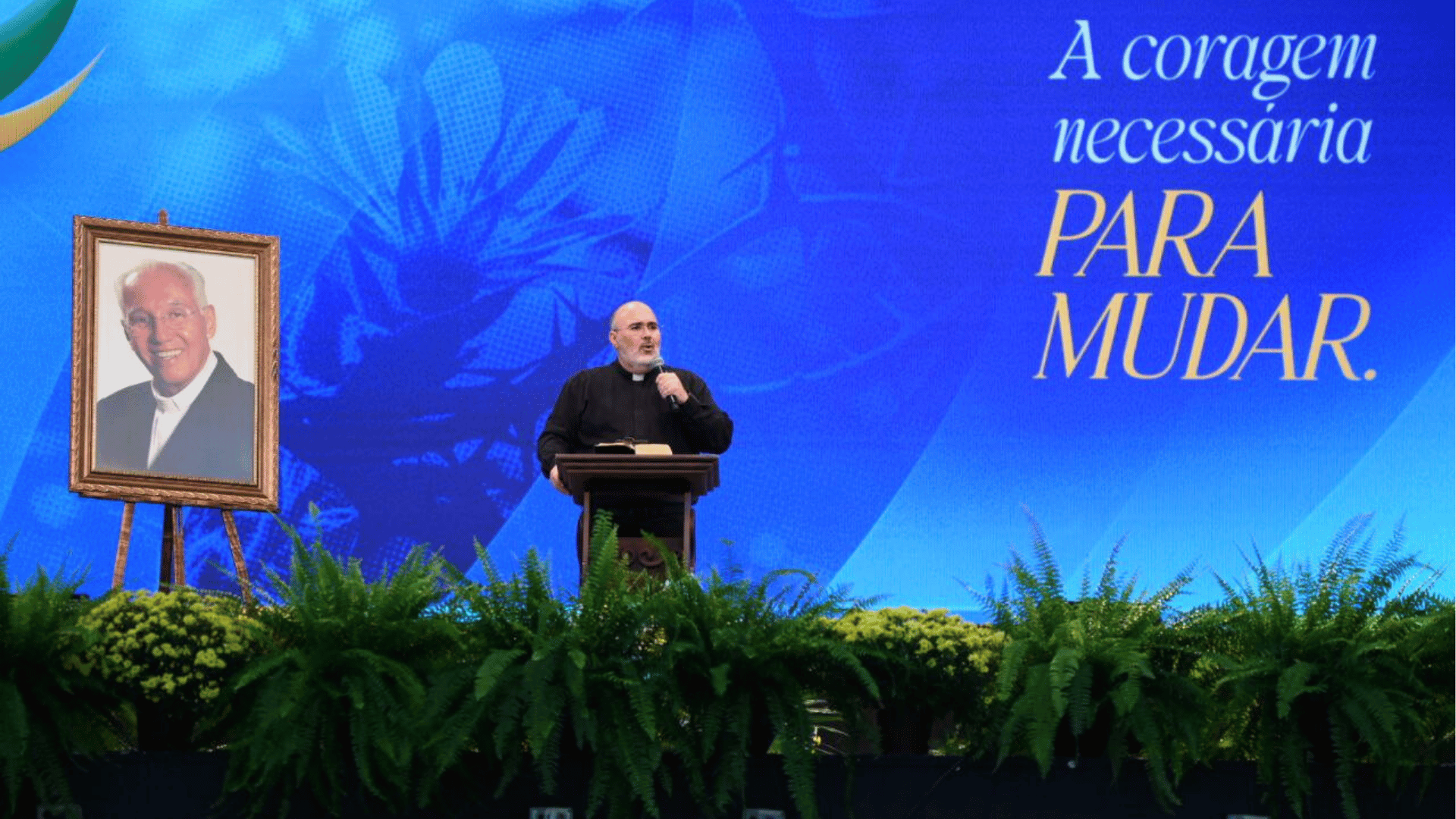 Padre Bruno Costa prega no palco do Hosana onde também há um quadro com imagem do padre Jonas Abib A imagem mostra um homem com vestes pretas e colarinho clerical - este homem é o padre Bruno Costa - em pé em um palco e ao seu lado há um quadro com imagem do padre Jonas Abib. O telão ao fundo o palco exibe o escrito "A coragem necessária para mudar"