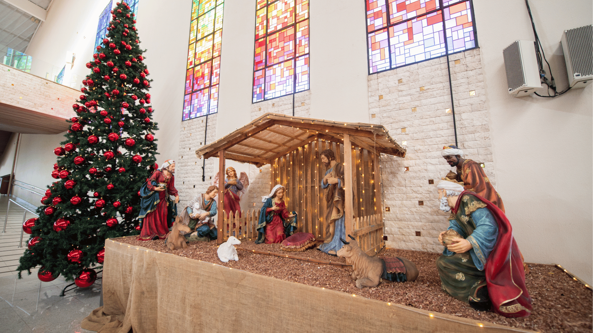 Presépio montado dentro de uma igreja, com figuras de Maria, José, o Menino Jesus, anjo, pastores, reis magos e animais em uma representação de um estábulo iluminado; ao lado, uma grande árvore de Natal decorada com bolas vermelhas, e ao fundo vitrais coloridos nas paredes.