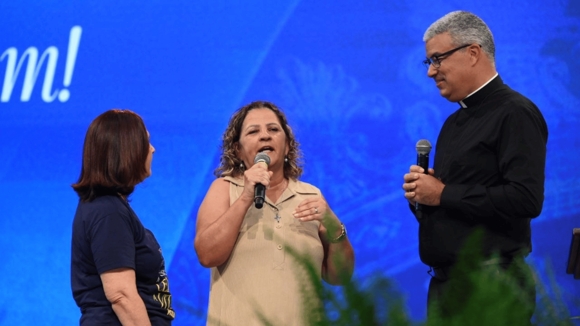 A imagem ilustra uma mulher de pele branca e cabelos cacheados loiros, usando um vestido de cor bege, com um microfone na mão, contando um testemunho. Duas pessoas a escutam: à esquerda, uma mulher de pele branca e cabelo castanho curto, usando uma camisa azul marinho, e à direita, um padre católico de pele branca e cabelos grisalhos, usando óculos e clérgima, também com um microfone nas mãos.