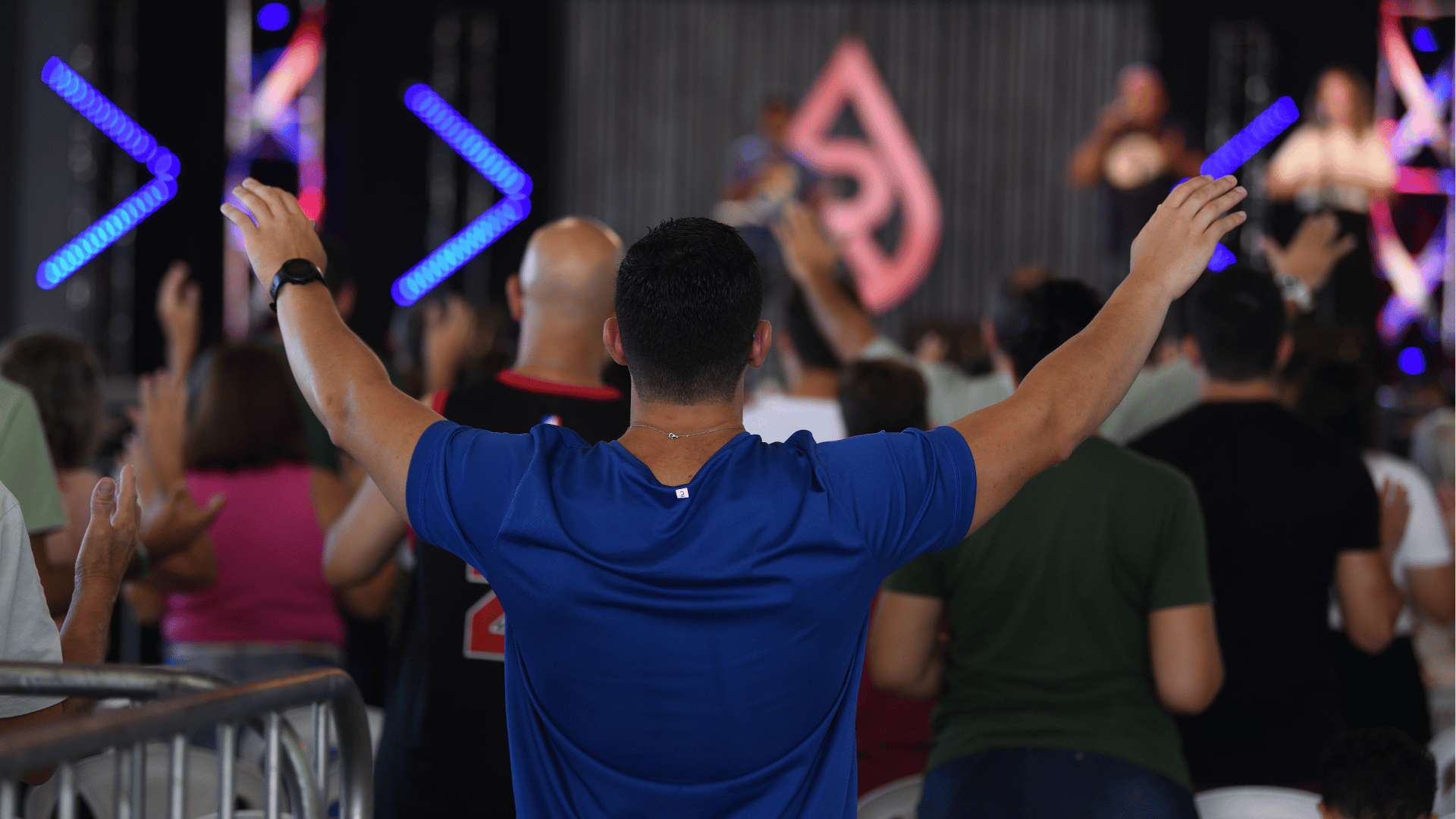 Um jovem de camiseta azul aparece de costas, com os braços elevados, em posição de louvor. À frente dele há outras pessoas  e o palco com a identidade do Revolução Jesus