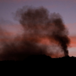Coluna de fumaça escura sobe de área montanhosa ao entardecer, contrastando com o céu em tons rosados.