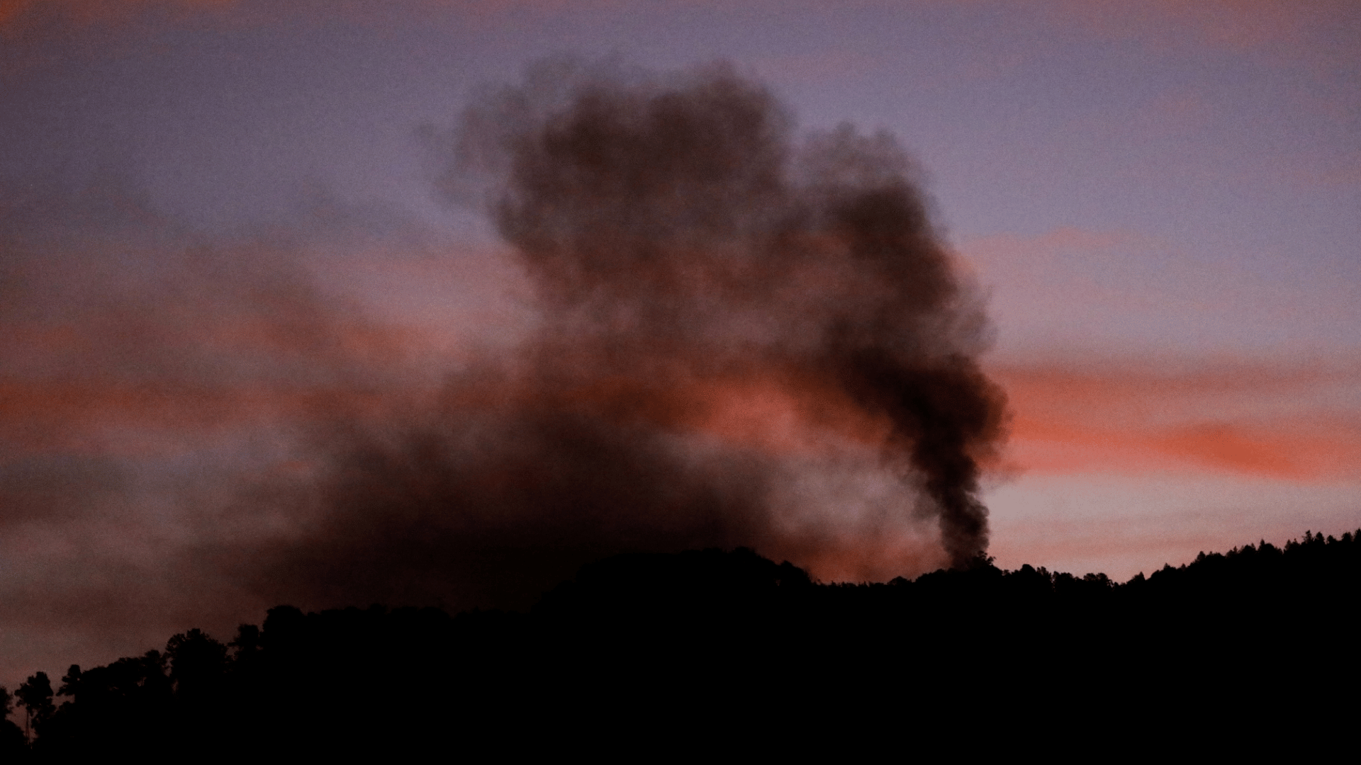 Coluna de fumaça escura sobe de área montanhosa ao entardecer, contrastando com o céu em tons rosados.