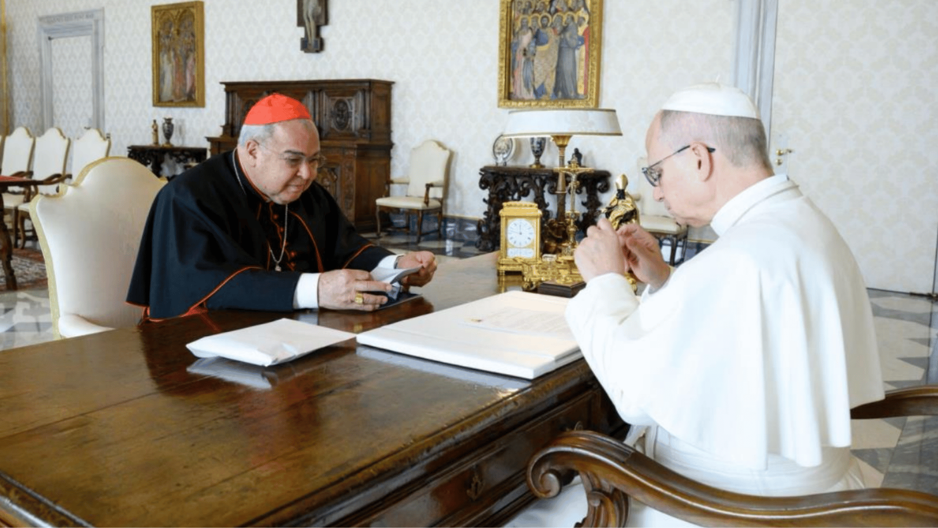 A imagem ilustra o Papa Leão XIV com suas habituais vestes pontifícias sentado diante de sua mesa em seu escritório. À sua frente, também sentado, está o Cardeal João Orani Tempesta, com as habituais vestes do cardinalato.