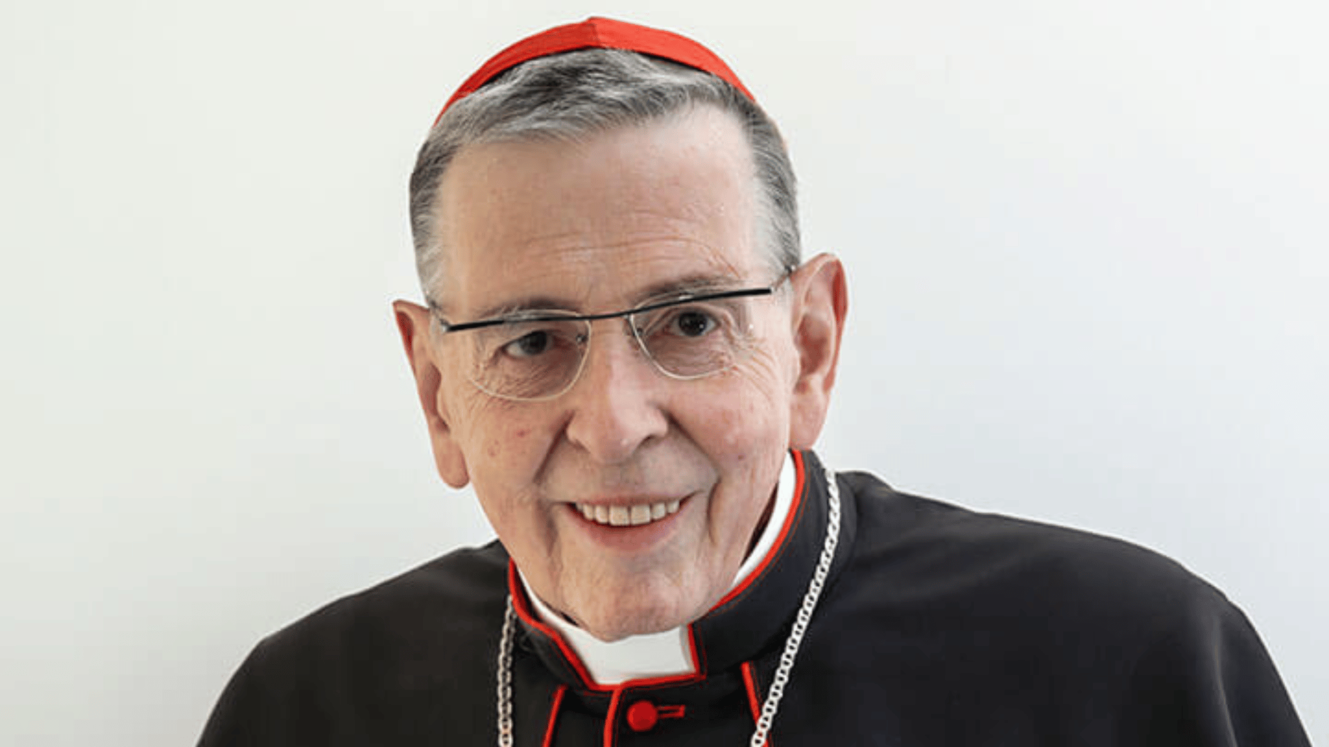 A imagem ilustra um cardeal da Igreja Católica, usando vestes pretas, clérgima e solidéu vermelho, de pele branca, cabelos grisalhos e óculos.