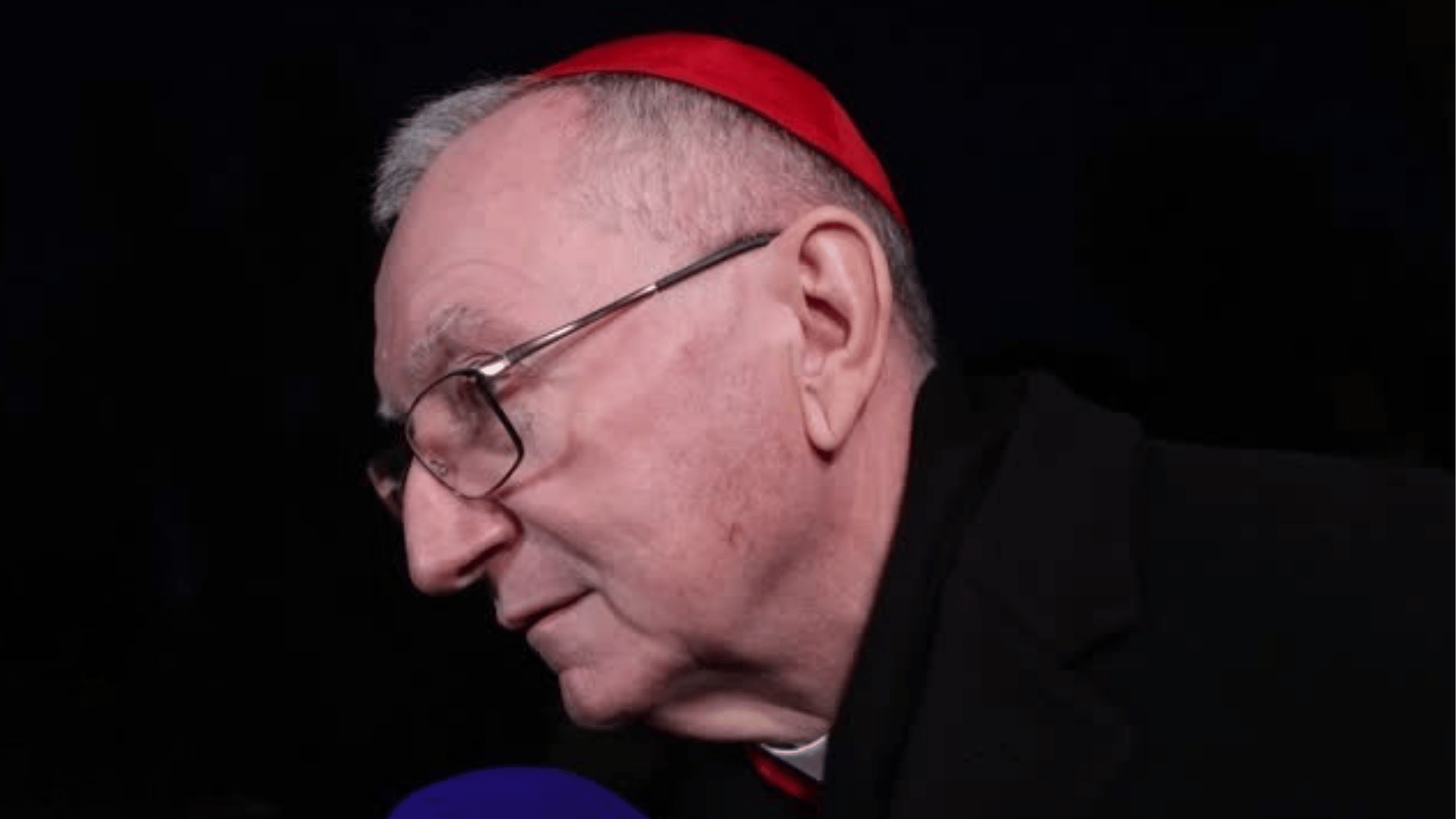 Cardeal de perfil, usando solidéu vermelho e óculos, falando ao microfone durante entrevista, com fundo escuro.