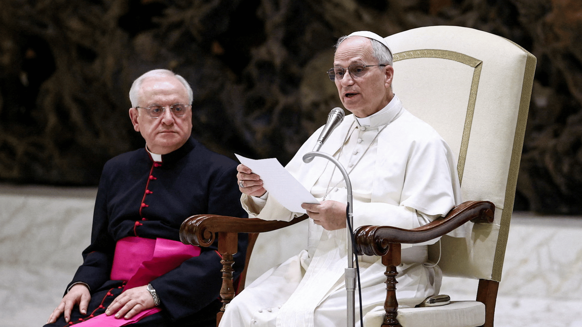 A imagem ilstra o Papa Leão XIV vestido com suas habituais vestes pontifícias, sentado em uma cadeira, segurando folhas de papel em suas mãos e com um microfone à sua frente. Ao seu lado direito está sentado um bispo de pele branca e cabelos grisalhos.