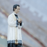 Imagem de Dom Bosco, sacerdote católico, com as mãos postas em oração, vestindo batina preta e sobrepeliz branca, em ambiente interno do Santuário do Pai das Misericórdias