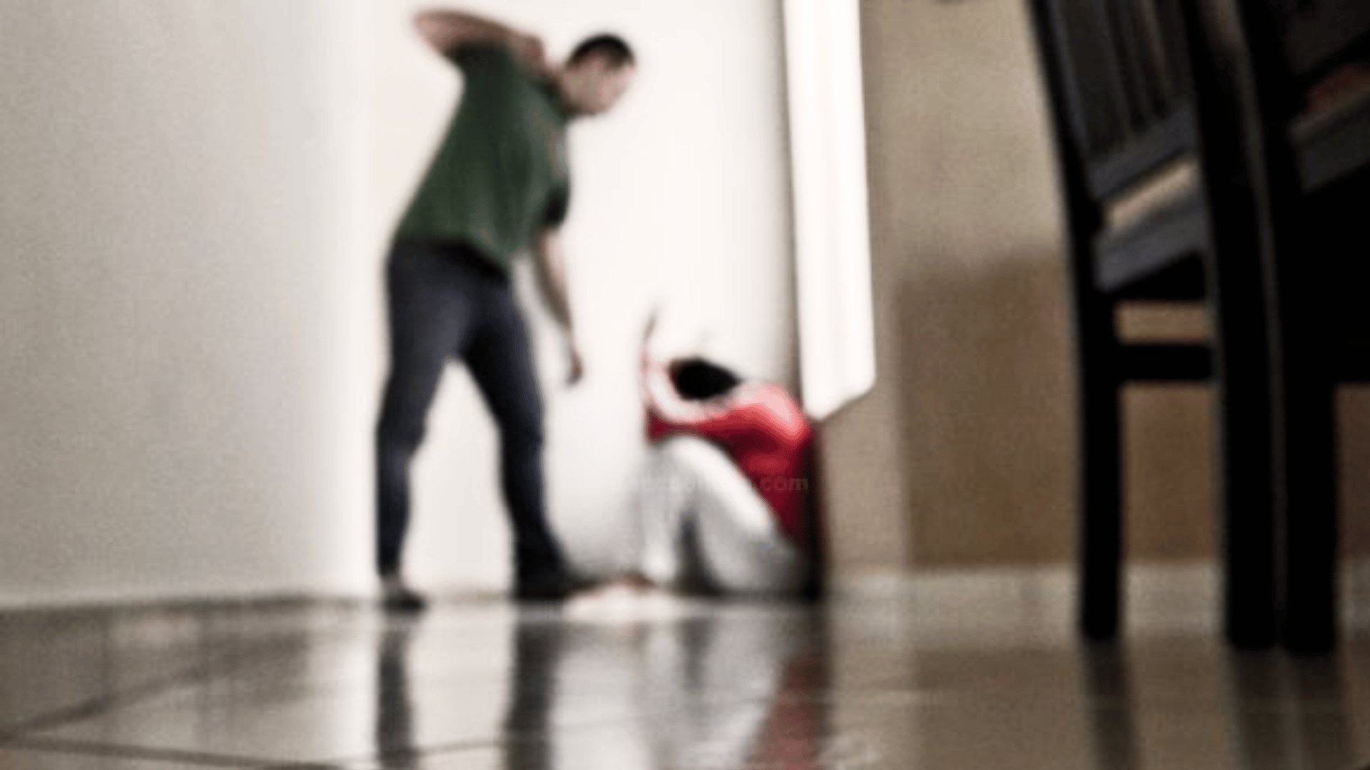 Imagem desfocada mostra um homem em pé com o braço levantado diante de uma mulher caída no chão, em ambiente interno, sugerindo situação de violência doméstica.