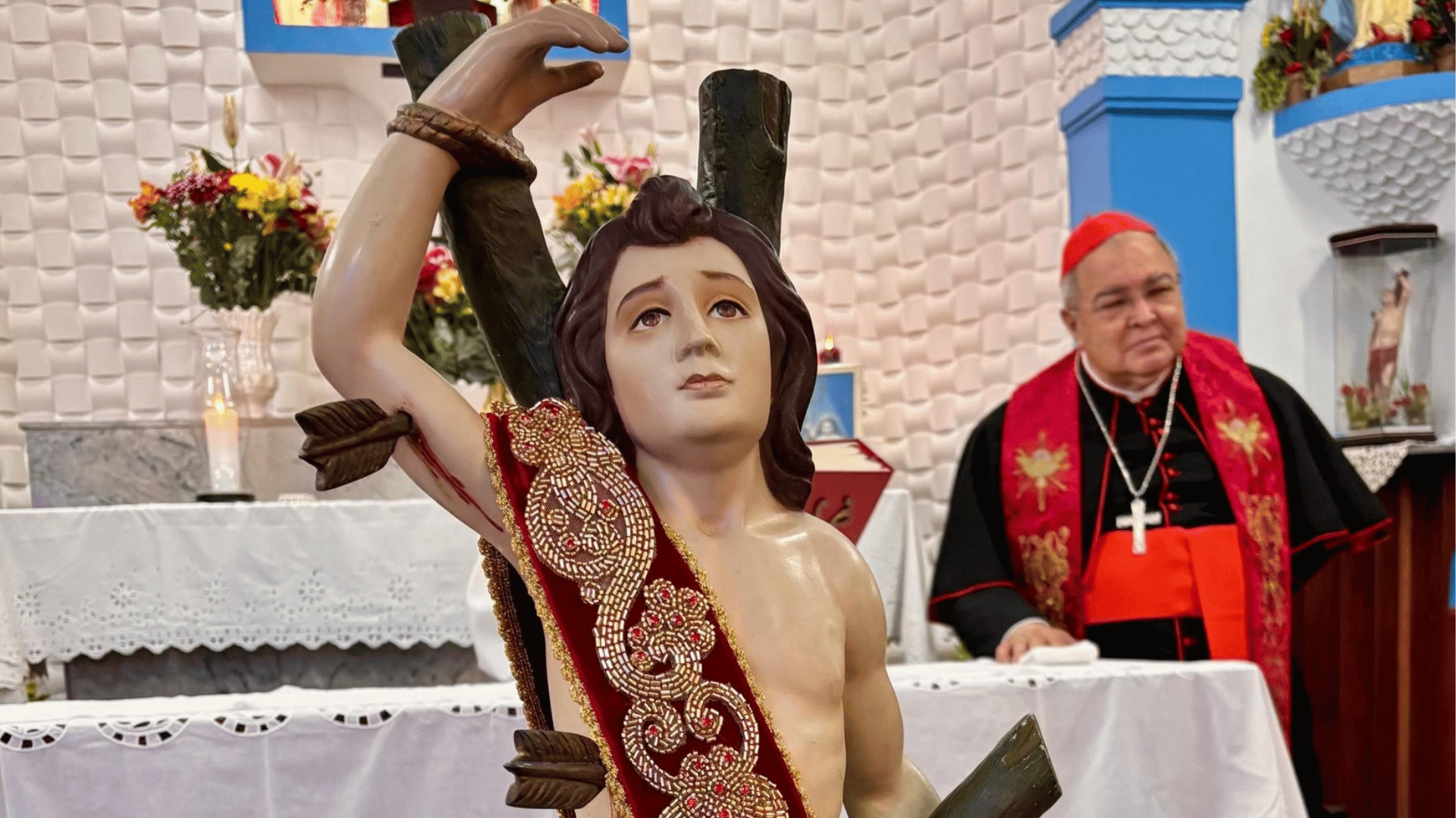 Imagem de São Sebastião amarrado a um tronco, com vestes vermelhas, diante do altar de uma igreja; ao fundo, o cardeal Orani Tempesta