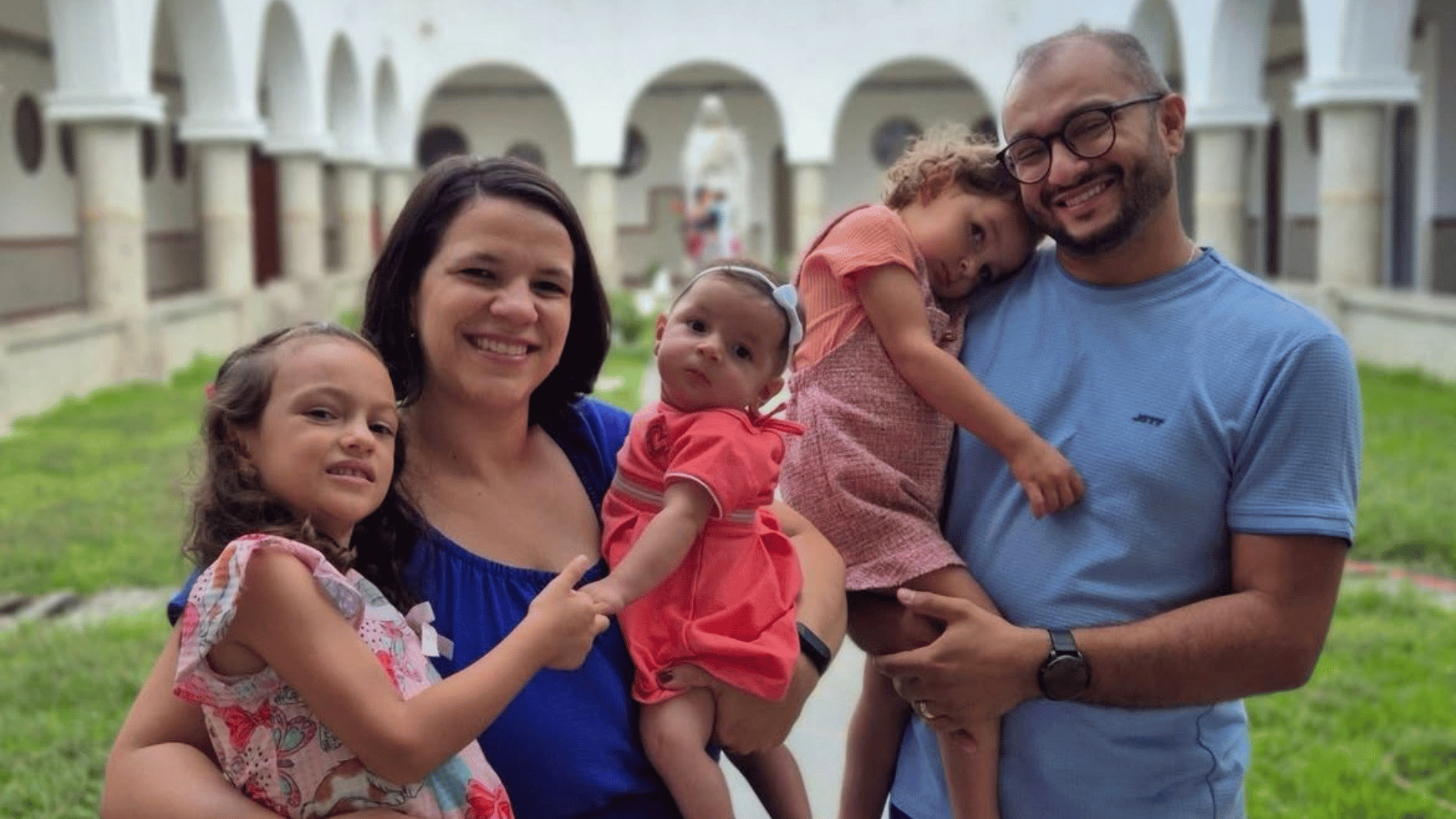 A imagem ilustra uma família composta por uma mãe, um pai e três filhas. A mãe segura a filha mais nova em um braço e abraça a mais velha com o outro enquanto o pai segura a filha do meio no colo.