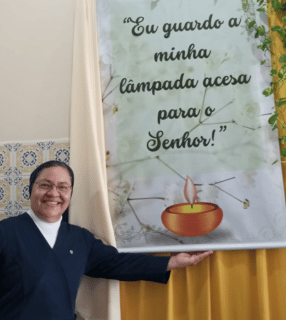 A imagem ilustra uma religiosa de pele branca e hábito azul, com o braço esquerdo estendido apontando para um banner no qual está escrito "Eu guardo a minha lâmpada acesa para o Senhor!".