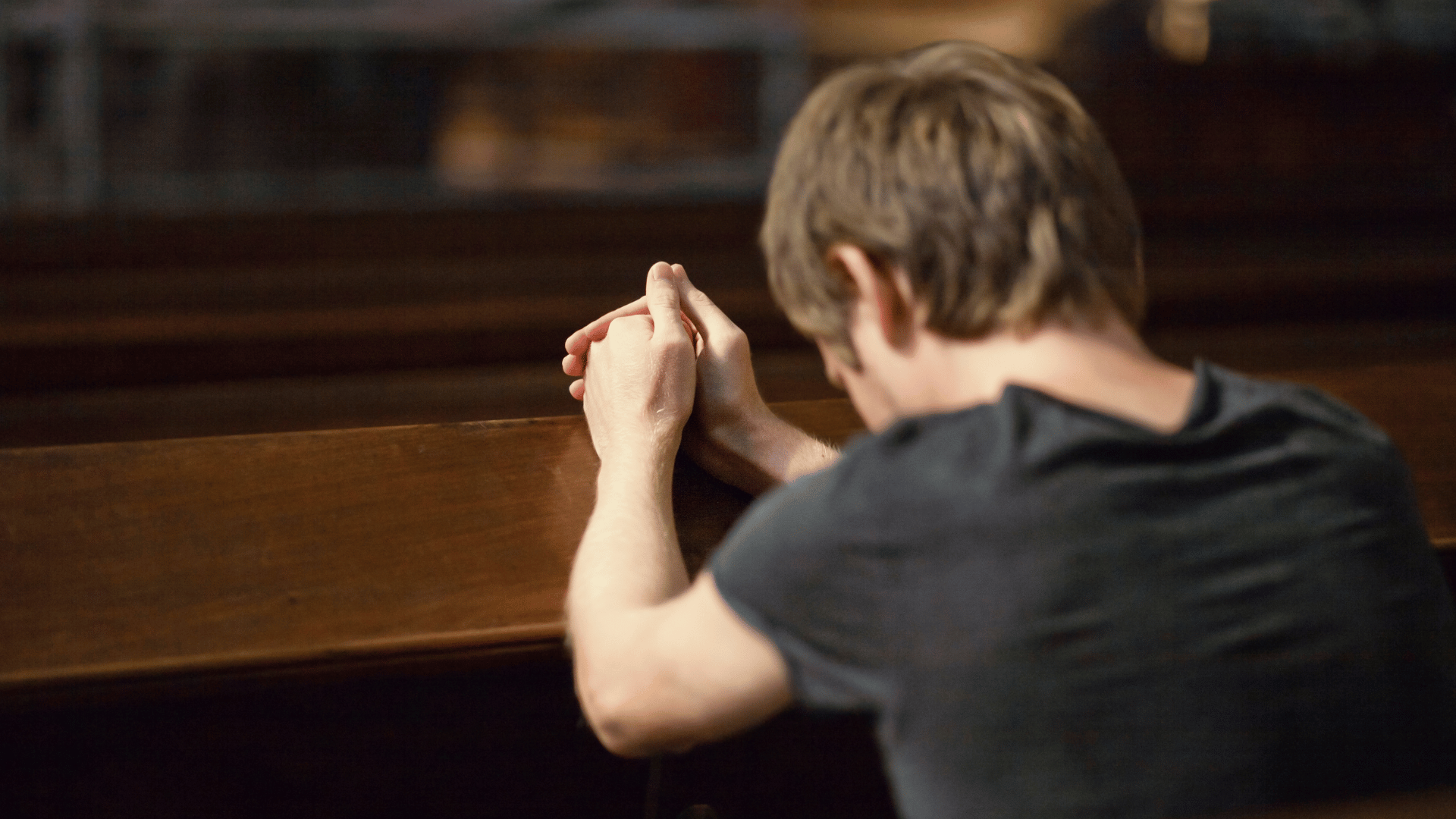 A imagem ilustra um homem branco, de cabelos curtos loiros e vestindo uma camisa preta ajoelhado diante de um banco de madeira, com as mãos unidas em gesto de oração.