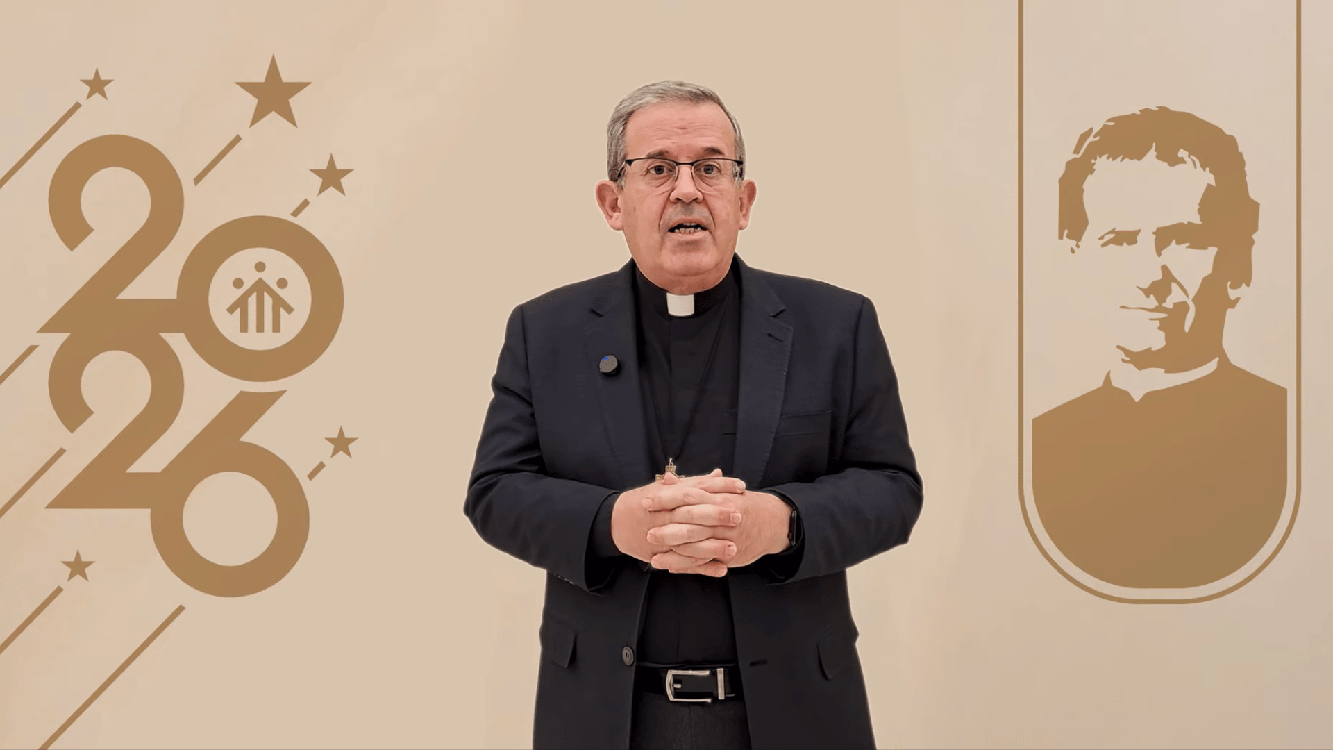 A imagem ilustra um sacerdote católico de pele branca e cabelos grisalhos. Ele usa um óculos, veste uma clérgima com um blazer e as mãos unidas sobre a barriga. O fundo da imagem é bege e inclui a inscrição "2026" estilizada do lado esquerdo e um ícone de Dom Bosco do lado direito.